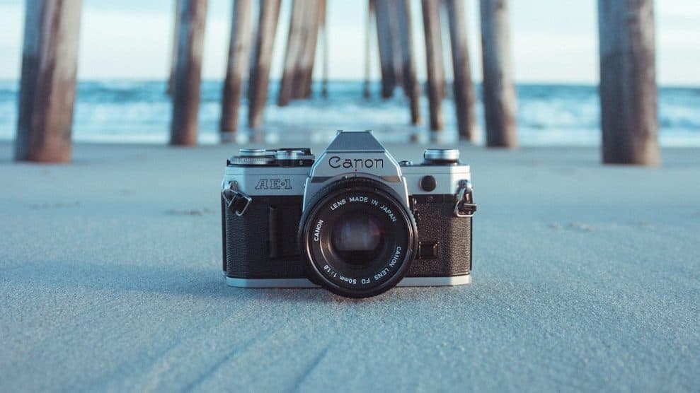 black and gray Canon AE-1 camera on gray sand under brown dock near body of water at daytime